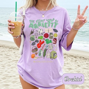 May include: A light purple t-shirt with the text "THE SECRET WORLD OF ARRIETTY" and various illustrated objects. The shirt features a graphic design with a variety of colorful illustrations. The word "Orchid" is written in a cursive font at the bottom right.