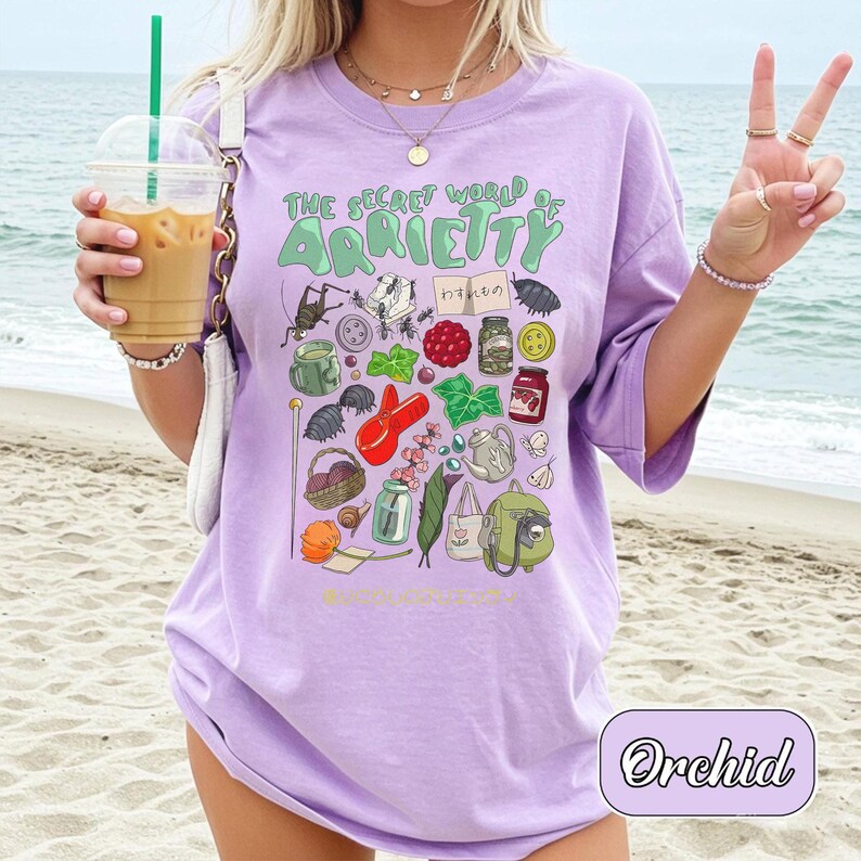 May include: A light purple t-shirt with the text "THE SECRET WORLD OF ARRIETTY" and various illustrated objects. The shirt features a graphic design with a variety of colorful illustrations. The word "Orchid" is written in a cursive font at the bottom right.