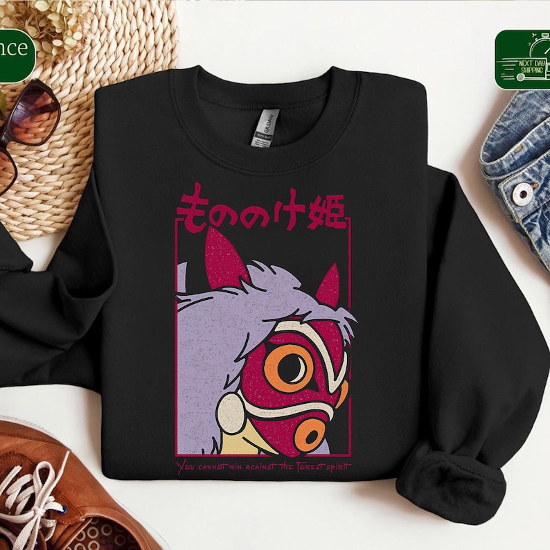 Puede incluir: Sudadera negra de cuello redondo con un gr&aacute;fico de un personaje de la pel&iacute;cula de animaci&oacute;n, La Princesa Mononoke. El dise&ntilde;o incluye una m&aacute;scara roja con detalles naranjas, pelo morado y texto japon&eacute;s. El texto "You cannot win against the forest spirit" est&aacute; en la parte inferior.