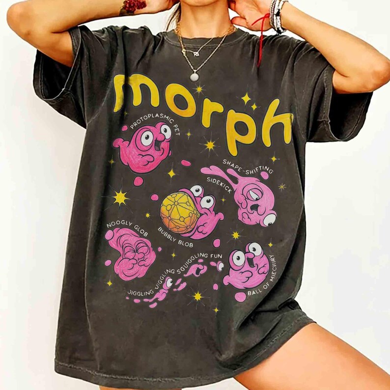 May include: A dark grey t-shirt with the word "Morph" in yellow, showcasing pink cartoon blob characters with labels such as "Noogly Glob" and "Bubbly Blob." The design incorporates stars and text.