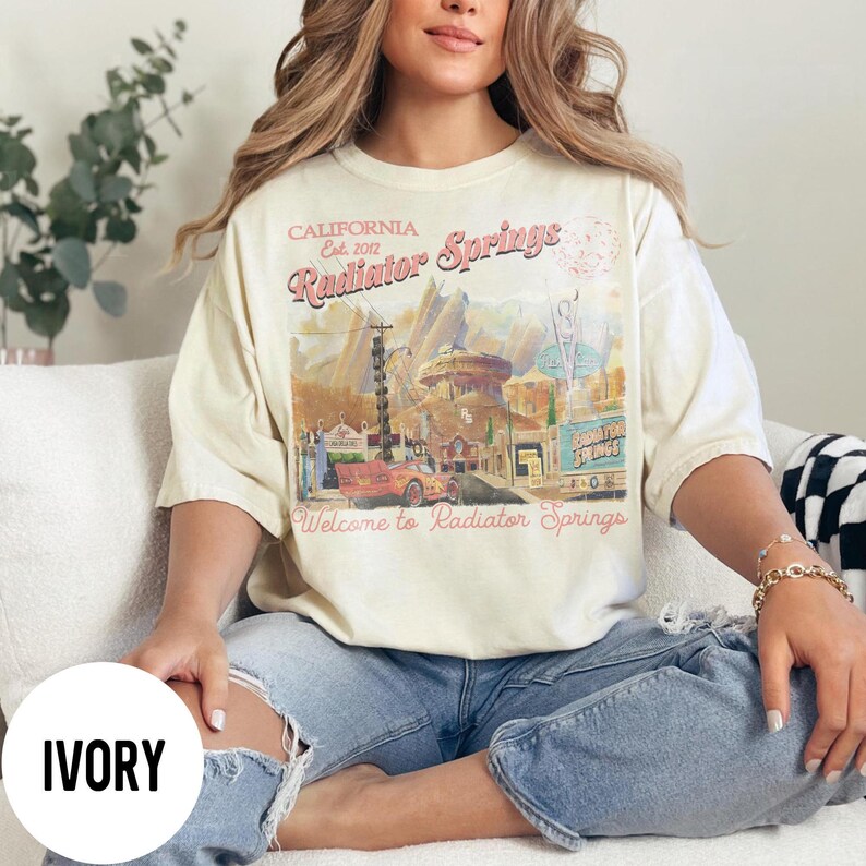 May include: Ivory-colored t-shirt featuring a graphic of Radiator Springs, California, with text that reads "Welcome to Radiator Springs." The design includes a red car and various buildings. The shirt is a relaxed fit.