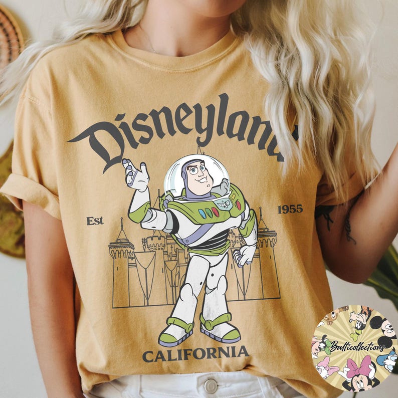 May include: Mustard yellow t-shirt featuring a graphic of Buzz Lightyear from Toy Story. The design includes the text "Disneyland California" and the year "1955". The shirt has rolled-up sleeves and a round neckline.