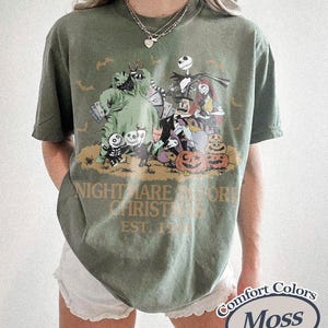 May include: A moss green short-sleeve t-shirt featuring a graphic print of characters from The Nightmare Before Christmas, including Jack Skellington, Sally, and Oogie Boogie, with the text Nightmare Before Christmas Est. 1993 printed below.