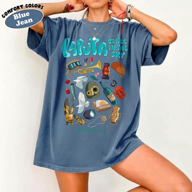 May include: Blue jean-colored t-shirt with a colorful graphic featuring the text "Laputa Castle in the Sky" and various illustrations. The design includes a trumpet, a bottle, and other themed elements.