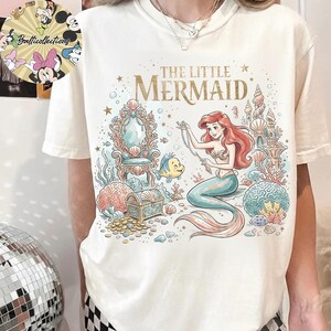 May include: A cream-colored t-shirt featuring a graphic print of The Little Mermaid with Ariel, Flounder, a treasure chest, and an ornate chair. The text reads THE LITTLE MERMAID in gold lettering. The design includes coral, seashells, and bubbles.