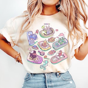 K&ouml;nnte beinhalten: Cremefarbenes T-Shirt mit einem farbenfrohen Cartoon-Design von Charakteren in Mobiltelefonen. Das Design umfasst Rosa, Blau, Gelb und Gr&uuml;n. Das Shirt besteht aus einem weichen Material.