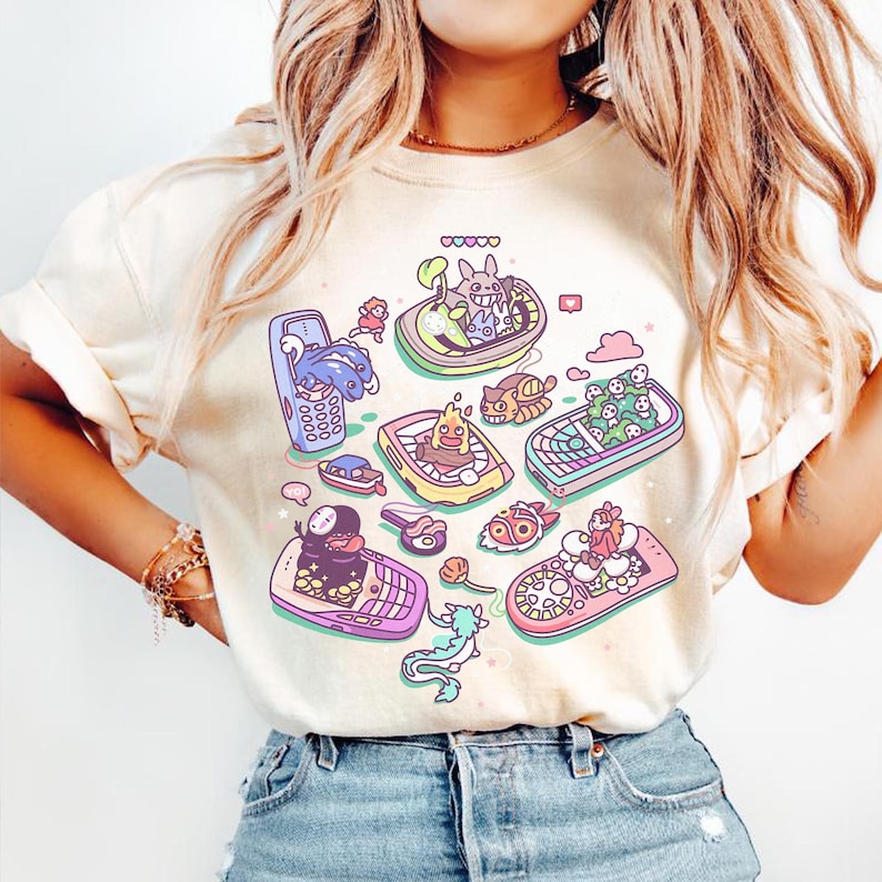 K&ouml;nnte beinhalten: Cremefarbenes T-Shirt mit einem farbenfrohen Cartoon-Design von Charakteren in Mobiltelefonen. Das Design umfasst Rosa, Blau, Gelb und Gr&uuml;n. Das Shirt besteht aus einem weichen Material.
