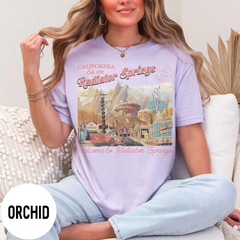 May include: A light purple t-shirt with a graphic of Radiator Springs, California. The design includes a vintage car, buildings, and the text "CALIFORNIA Est. 2012 Radiator Springs Welcome to Radiator Springs."