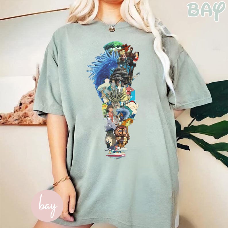 May include: Sage green t-shirt featuring a vertical collage of anime characters and scenes. The graphic includes various characters in a colorful arrangement. The shirt has short sleeves and a relaxed fit, with the word "bay" in a light green color.