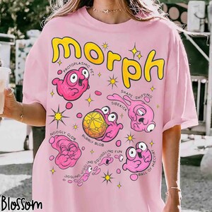 May include: Pale pink t-shirt with the word "Morph" in yellow bubble letters. The shirt features cartoon illustrations of pink blob creatures with labels such as "Protoplasmic Pet" and "Shape-shifting Sidekick".