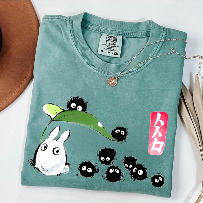 May include: A teal Comfort Colors t-shirt featuring a white rabbit-like character with a green leaf and several black soot sprites. The shirt also has Japanese text on a red rectangle and a Comfort Colors tag.