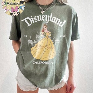 May include: Olive green t-shirt featuring a Disneyland graphic. The design includes the word "Disneyland" in a stylized font, the image of Belle in a yellow dress, and the text "CALIFORNIA" and "Est 1955".