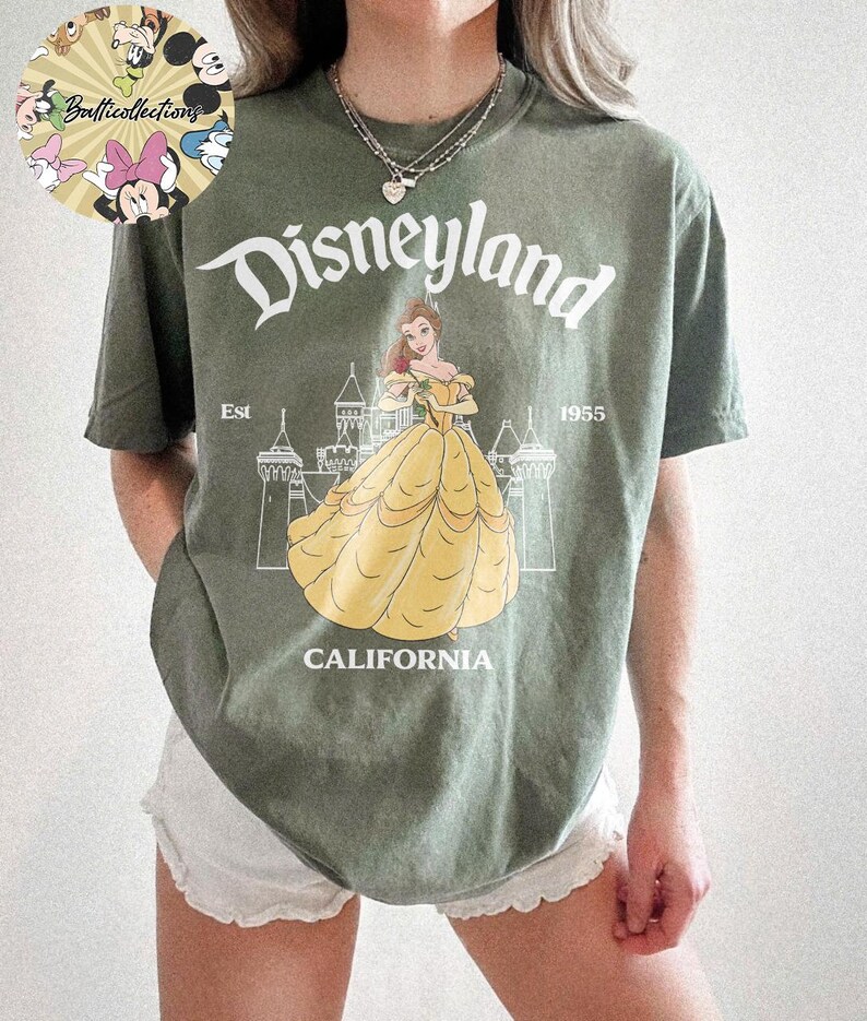 May include: Olive green t-shirt featuring a Disneyland graphic. The design includes the word "Disneyland" in a stylized font, the image of Belle in a yellow dress, and the text "CALIFORNIA" and "Est 1955".