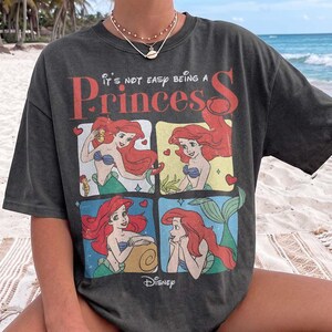 May include: A charcoal gray short-sleeve t-shirt featuring a graphic print of the character Ariel from The Little Mermaid in four distinct panels. The text reads It's NOT EASY BEING A Princess and includes the Disney logo at the bottom.