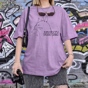 May include: Orchid-colored t-shirt with a black graphic of a cartoon character and Japanese text. The text "Studio Ghibli" is below the character. The shirt has short sleeves and a relaxed fit. The words "Orchid Comfort Colors" are in the upper left corner.