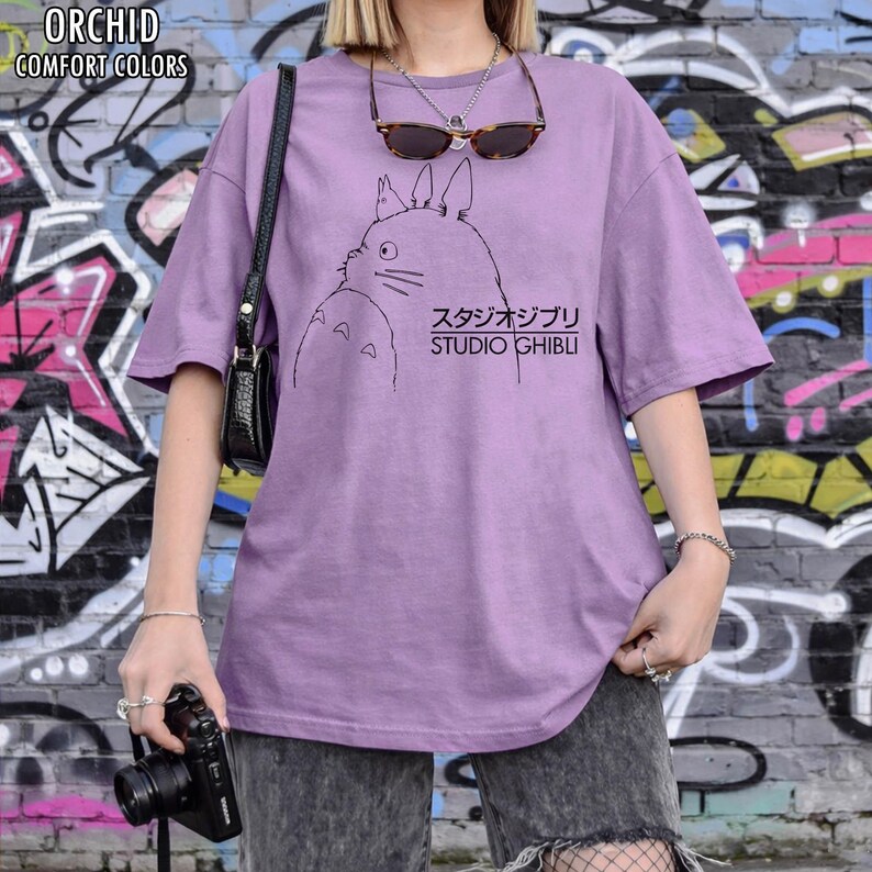 May include: Orchid-colored t-shirt with a black graphic of a cartoon character and Japanese text. The text "Studio Ghibli" is below the character. The shirt has short sleeves and a relaxed fit. The words "Orchid Comfort Colors" are in the upper left corner.