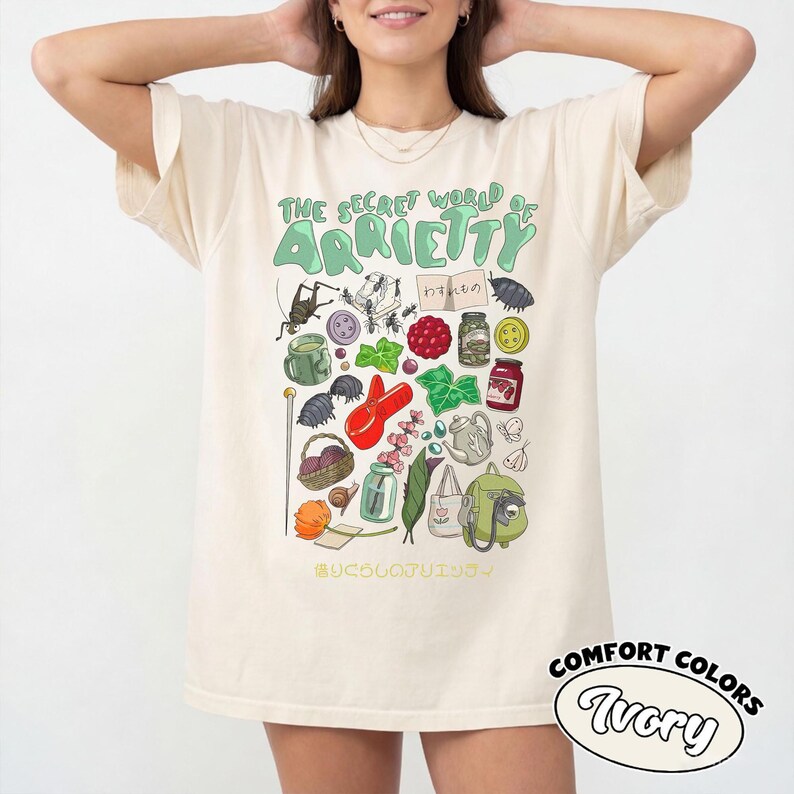 May include: Ivory-colored t-shirt featuring a whimsical illustration with the text "The Secret World of Arrietty." The design includes various small objects and Japanese text, with the words "Comfort Colors" and "Ivory" in a circular logo.
