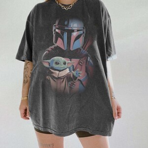 May include: A charcoal grey short-sleeve t-shirt featuring a graphic print of a helmeted warrior in silver and blue armor standing behind a small green creature with large ears and eyes, wearing a tan robe and reaching out with one hand.