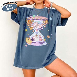 May include: Blue jean colored t-shirt with a whimsical hourglass design. The graphic features cartoon characters, flowers, stars, and a crescent moon. The shirt has the words "COMFORT COLORS" and "Blue Jean" in a circular label.