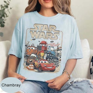 Puede incluir: Camiseta azul claro con un gr&aacute;fico de personajes de Cars y el logo de Star Wars. El dise&ntilde;o incluye a Rayo McQueen y otros veh&iacute;culos animados. La camiseta tiene la palabra "Chambray" en la parte inferior.