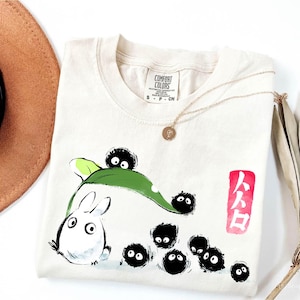 May include: Cream-colored t-shirt featuring a cartoon illustration of a white rabbit-like creature with a green leaf and several black soot sprites. The shirt also has Japanese text in a red box and a Comfort Colors tag.