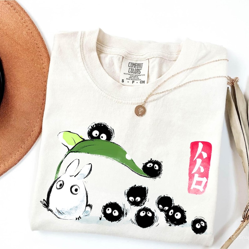 May include: Cream-colored t-shirt featuring a cartoon illustration of a white rabbit-like creature with a green leaf and several black soot sprites. The shirt also has Japanese text in a red box and a Comfort Colors tag.