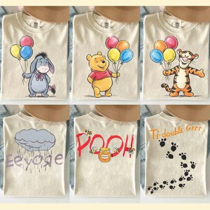 Könnte beinhalten: Cremefarbene T-Shirts mit verschiedenen Winnie-the-Pooh-Charakteren und -Designs. I-Aah, Puuh und Tiger halten Ballons. Andere Shirts zeigen Text und Pfotenabdrücke. Die Shirts liegen auf einer Jeansoberfläche.