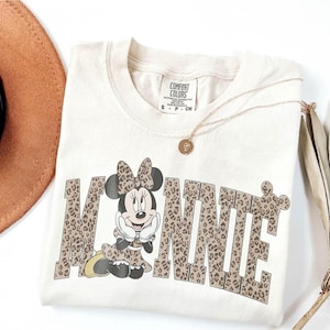 Puede incluir: Camiseta color crema con un gr&aacute;fico de Minnie Mouse. La palabra "MINNIE" est&aacute; impresa con un estampado de leopardo. Tambi&eacute;n se ven un collar dorado con un colgante y un sombrero marr&oacute;n.