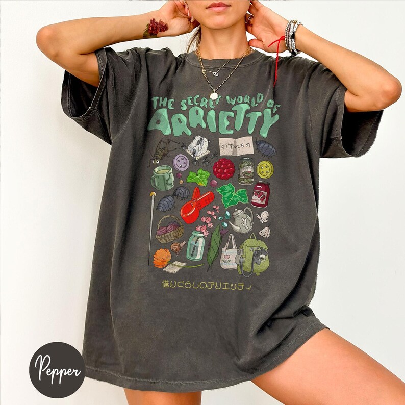 May include: Dark gray t-shirt featuring the text "THE SECRET WORLD OF ARRIETTY" in green, with various illustrated objects. The design includes a bug, buttons, berries, and other whimsical elements. The shirt has a relaxed fit.