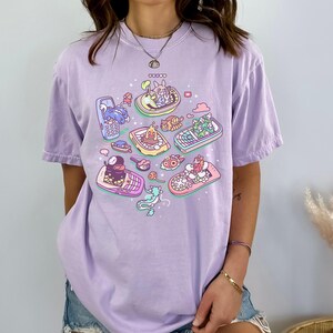 K&ouml;nnte beinhalten: Helllila T-Shirt mit einem farbenfrohen Grafikdesign von Zeichentrickfiguren in Retro-Handys. Das Design zeigt Charaktere in verschiedenfarbigen Handys und erzeugt eine skurrile und verspielte &Auml;sthetik.