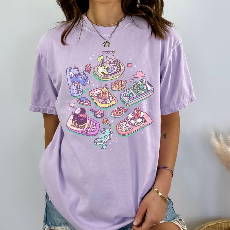 K&ouml;nnte beinhalten: Helllila T-Shirt mit einem farbenfrohen Grafikdesign von Zeichentrickfiguren in Retro-Handys. Das Design zeigt Charaktere in verschiedenfarbigen Handys und erzeugt eine skurrile und verspielte &Auml;sthetik.
