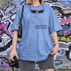 May include: A light blue t-shirt featuring a black outline illustration of a cartoon character and the text "STUDIO GHIBLI" in English and Japanese. The shirt also has the text "BLUE JEAN COMFORT COLORS" at the top.