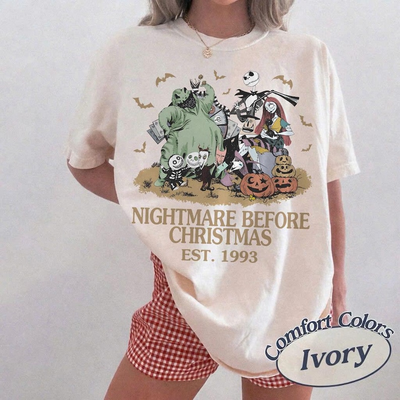 May include: An ivory short-sleeve t-shirt featuring a graphic print of characters from The Nightmare Before Christmas, including Jack Skellington, Sally, and Oogie Boogie. The text reads NIGHTMARE BEFORE CHRISTMAS EST. 1993 with small bats.