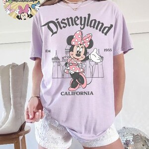 May include: A light purple t-shirt featuring a graphic of Minnie Mouse in a pink polka dot dress and bow, with the text "Disneyland California Est. 1955". The shirt is a relaxed fit.