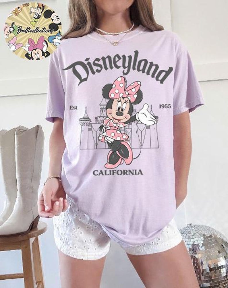 May include: A light purple t-shirt featuring a graphic of Minnie Mouse in a pink polka dot dress and bow, with the text "Disneyland California Est. 1955". The shirt is a relaxed fit.