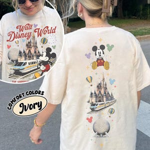 Pu&ograve; includere: Una t-shirt color avorio con una stampa grafica verticale di Topolino, un castello, una monorotaia e una cupola geodetica, arricchita da palloncini e stelle colorati. Il testo Walt Disney World &egrave; stampato in rosso sopra l'illustrazione del castello.