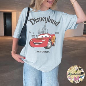 May include: Light blue t-shirt featuring a Disneyland graphic. The design includes the Disneyland logo, a cartoon car, a castle, and the text "CALIFORNIA" at the bottom. The shirt is a relaxed fit.