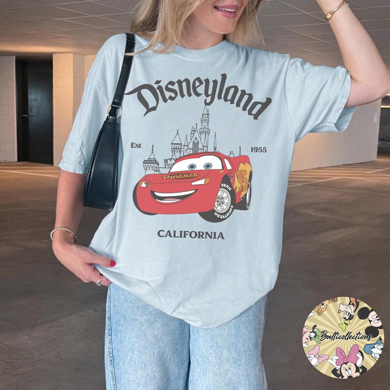 May include: Light blue t-shirt featuring a Disneyland graphic. The design includes the Disneyland logo, a cartoon car, a castle, and the text "CALIFORNIA" at the bottom. The shirt is a relaxed fit.