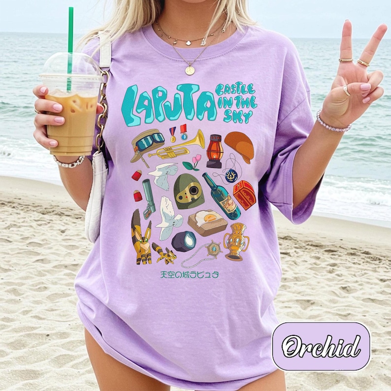 May include: A light purple t-shirt with the words "LAPUTA" and "CASTLE IN THE SKY" in a teal font. The shirt features various cartoon illustrations, including a trumpet, a bottle, and a trophy. The word "Orchid" is in a white cursive font at the bottom.