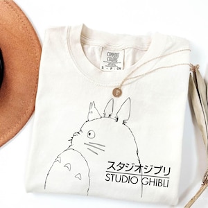 May include: Cream-colored t-shirt featuring a black graphic of a cartoon character with the text "STUDIO GHIBLI" in Japanese and English. A gold necklace with a pendant rests on the shirt. A brown hat is visible in the upper left corner.