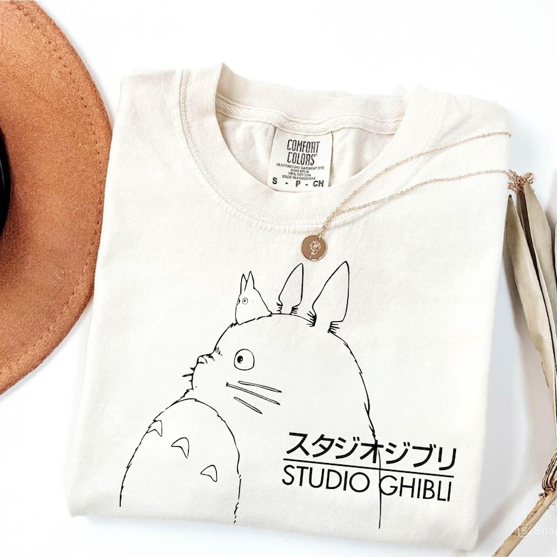 May include: Cream-colored t-shirt featuring a black graphic of a cartoon character with the text "STUDIO GHIBLI" in Japanese and English. A gold necklace with a pendant rests on the shirt. A brown hat is visible in the upper left corner.