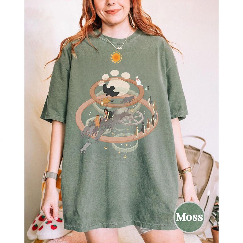 May include: Moss green t-shirt featuring a whimsical cat-themed graphic. The design includes a central circular arrangement with cats, a person, and a paw print. The shirt has a relaxed fit and short sleeves.