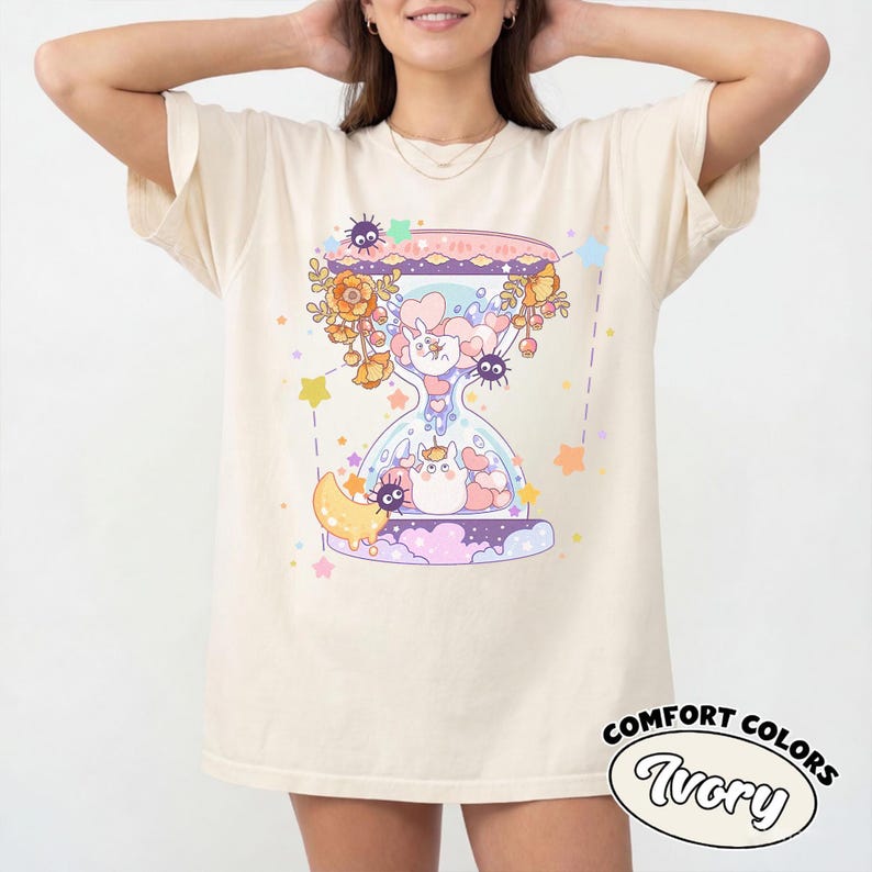 May include: Ivory-colored t-shirt featuring a whimsical hourglass design with cartoon characters, flowers, and stars. The hourglass contains a unicorn and other creatures. The text "COMFORT COLORS Ivory" is printed on the lower right.