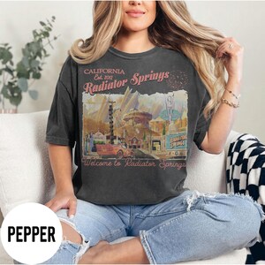 May include: Dark gray t-shirt featuring a vintage-style illustration of Radiator Springs, with text that reads "CALIFORNIA Est. 2012 Radiator Springs" and "Welcome to Radiator Springs". The design includes a red car and desert landscape.