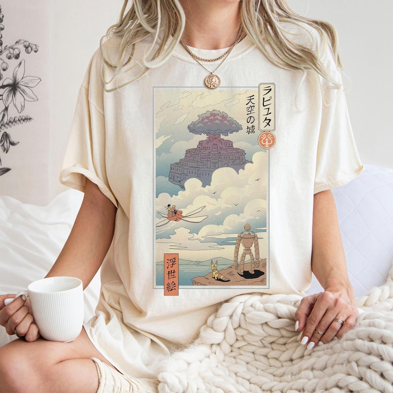 May include: Cream-colored t-shirt featuring a Japanese anime-style illustration. The artwork depicts a floating castle in the clouds, with characters and Japanese text. The shirt has short sleeves and a relaxed fit.