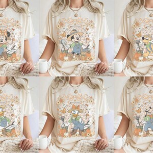 Puede incluir: Una camiseta de color crema con un estampado gráfico de varios personajes de Disney vestidos con ropa de jardinería. El diseño incluye calabazas, flores y herramientas de jardín enmarcadas por un lazo, mostrando un personaje distinto.