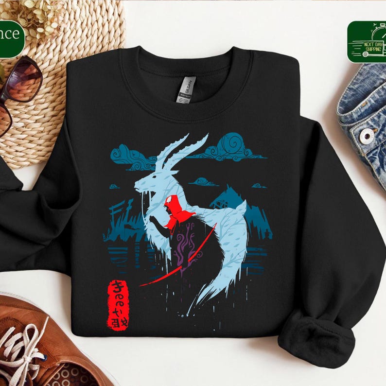Może przedstawiać: Czarna bluza z fantastyczną ilustracją. Przedstawia mityczne stworzenie z czerwoną postacią, niebieskimi chmurami i sylwetkami g&oacute;r. Obecny jest r&oacute;wnież japoński tekst.