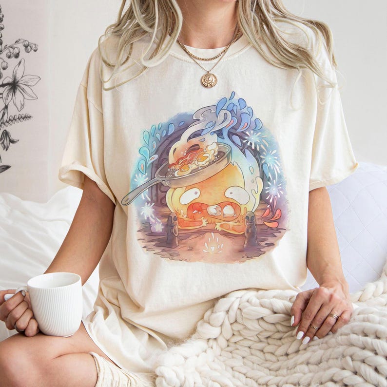 Puede incluir: Camiseta color crema con un personaje de huevo de dibujos animados cocinando huevos y tocino en una sart&eacute;n. El gr&aacute;fico utiliza colores azul, naranja y amarillo, con un estilo vintage desgastado. La camiseta tiene un corte holgado.