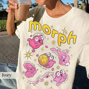 May include: Cream-coloured t-shirt with the word "Morph" in yellow, featuring cartoon pink blob creatures and text like "Protoplasmic Pet" and "Shape Shifting". The shirt has a crew neck.