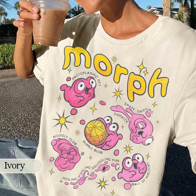 May include: Cream-coloured t-shirt with the word "Morph" in yellow, featuring cartoon pink blob creatures and text like "Protoplasmic Pet" and "Shape Shifting". The shirt has a crew neck.